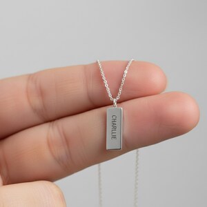 May include: A silver-colored necklace with a rectangular pendant. The pendant has the name "CHARLIE" engraved vertically. The necklace is held by a hand against a light gray background.