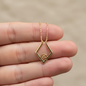May include: A gold necklace with a diamond-shaped pendant and a floral motif. The pendant hangs from a fine chain. The necklace is held in a hand against a blurred background. The pendant's design is a small flower.