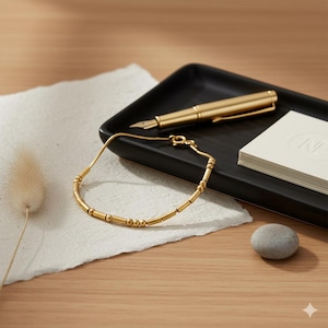 May include: A gold bracelet with cylindrical and spherical beads rests on a textured white paper. A gold fountain pen and a stack of white cards are on a black tray. A small gray stone and dried plant are also present.