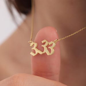 Può includere: Una collana color oro con un ciondolo con il numero "333". Il ciondolo è in un carattere stilizzato ed è sospeso a una catena delicata. La collana è mostrata su uno sfondo sfocato.