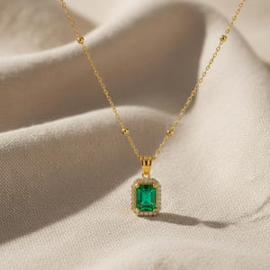 Könnte beinhalten: Eine goldfarbene Halskette mit einem rechteckigen, smaragdgrünen Anhänger. Der Anhänger ist von kleinen, klaren Edelsteinen umgeben. Die Kette hat kleine goldene Perlen und ein filigranes Design. Der Schmuck wird auf einem neutralfarbenen Stoff präsentiert.