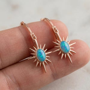 May include: Rose gold earrings featuring a starburst design with a central oval-shaped blue opal stone. The earrings have a lever back closure. The opal has shades of blue and green.
