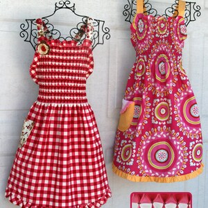 May include: Two aprons hang on decorative metal hangers. The apron on the left is red and white gingham with a honeycomb smocked bodice. The apron on the right is pink with a floral pattern. The aprons are labeled "Honeycomb Smocked Apron" and "Ladies Sm/Med/Lg."