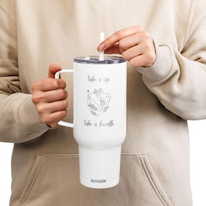 Könnte beinhalten: Weißer Becher mit Henkel und Strohhalm. Der Becher hat die Aufschrift "take a sip, take a breath" und ein herzförmiges Design mit Händen und Blättern. Der Markenname "koozie" steht unten.