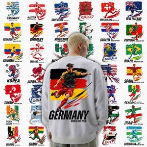 Op de afbeelding: Wit sweatshirt met een voetballer en het woord "GERMANY" in zwart, met "WORLD CUP 2026" eronder. Het ontwerp bevat vlaggen en voetballers die verschillende landen vertegenwoordigen.