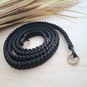 Könnte beinhalten: Ein schwarzer Paracord-Gurt mit goldfarbener Metallhardware. Der Gurt ist eng gewebt und aufgerollt, mit einem kreisförmigen Verschluss an einem Ende und einem kleineren Verschluss am anderen. Der Hintergrund ist eine helle Holzoberfläche.