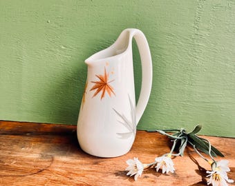 May include: White ceramic pitcher with a curved handle and a spout. The pitcher is decorated with orange and gray leaf designs. The pitcher is on a wooden surface with white flowers and green leaves.