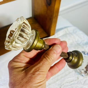 May include: A vintage-style door knob set featuring two clear, faceted glass knobs with an octagonal shape. The knobs are connected by a central metal spindle and are mounted on circular, aged brass-toned metal bases with a weathered finish.