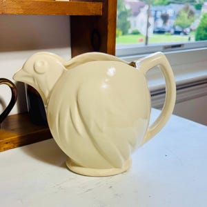 May include: A cream-colored ceramic pitcher shaped like a bird, featuring a molded beak and eye on the left side, a rounded body with embossed wing details, a circular base, and a sturdy handle on the right side for pouring liquids.