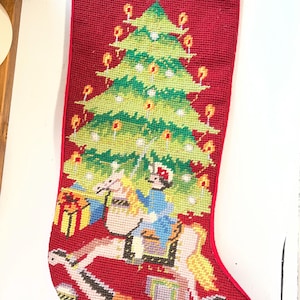 May include: A red needlepoint Christmas stocking featuring a large green Christmas tree with yellow candles, a figure riding a white rocking horse, and wrapped gifts. The stocking has a cream-colored cuff at the top and a red hanging loop.