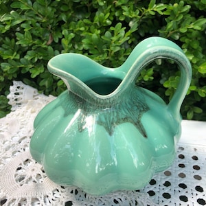 May include: A light green ceramic pitcher with a rounded, bulbous body and a flared spout. The pitcher has a textured, slightly raised design around the top and a handle that curves back towards the spout.
