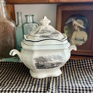 May include: A white ceramic tureen with a matching lid, featuring black and white landscape illustrations on both the body and the lid. The tureen has two side handles, a scalloped rim, and a decorative finial on top of the lid.
