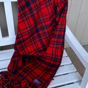 May include: A red and navy blue plaid blanket featuring a woven pattern of intersecting lines. The blanket has a rectangular shape with fringed edges at the bottom and a small blue rectangular label with white text attached to the lower corner.