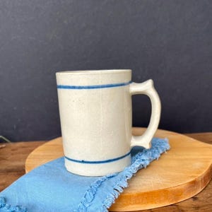 May include: A white ceramic mug with blue stripes around the top and bottom. The mug has a handle and is sitting on a wooden surface.