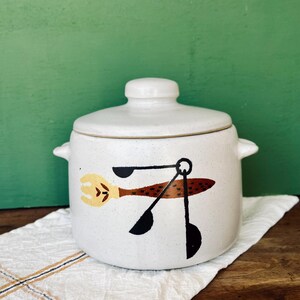 May include: White ceramic crock with a lid, featuring a brown and black stylized design of kitchen utensils, including a fork, spoon, and ladle.