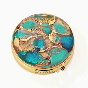 May include: A round, gold-toned compact mirror with a teal, blue, and gold marbled design. The mirror has a hinged lid and a small, round, gold-toned clasp.