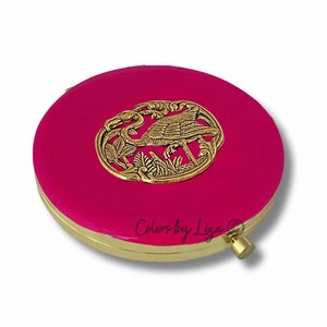 May include: A round, pink compact mirror with a gold metal design of a bird in a floral frame. The mirror has a hinged lid and a small gold knob on the side.
