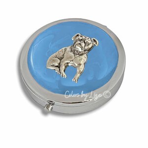 May include: A silver compact mirror with a blue background and a silver bulldog image. The mirror has a hinged lid and a small latch.