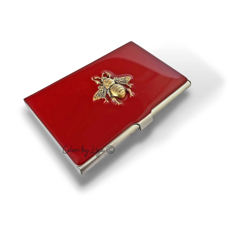 Puede incluir: Porta tarjetas de visita de metal rojo con un dise&ntilde;o de abeja dorada. El porta tarjetas tiene un borde plateado y el texto "Colors by Liga &copy;" est&aacute; impreso en la parte inferior.