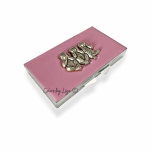 May include: A rectangular, pink pill box with a silver-colored metal trim. The lid features three silver monkey figurines in the "See no evil, hear no evil, speak no evil" pose. The text "Colors by Liga" is visible on the bottom left.