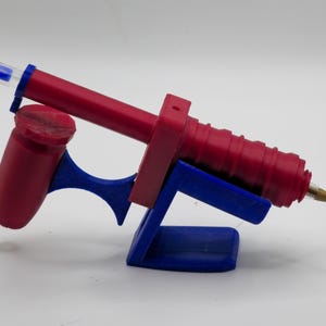 May include: A red and blue glue gun with a blue stand. The glue gun has a red barrel and handle, with a blue trigger and stand. The tip of the glue gun is gold. The glue gun is designed for crafting and DIY projects.