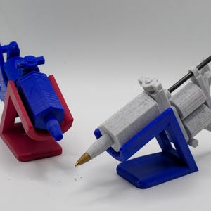 Puede incluir: Dos soportes para máquinas de tatuaje impresos en 3D. Un soporte es rojo y el otro es azul. Las máquinas de tatuaje son grises y azules.