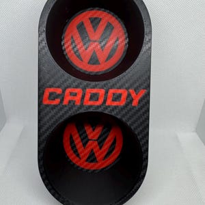 May include: Black double cup holder with a carbon fibre pattern. The cup holder has two red Volkswagen logos and the word "CADDY" in red letters.