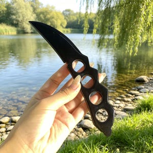 Könnte beinhalten: Ein durchgehend schwarzes Messer mit glatter Oberfläche, das eine geschwungene Klinge und einen Griff mit vier integrierten kreisförmigen Fingerlöchern aufweist. Das Objekt besteht aus einem dunklen, polierten Material und wirkt einheitlich.