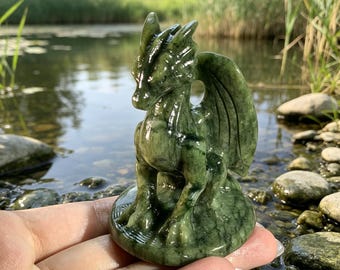 May include: A carved green stone dragon figurine featuring a smooth, polished surface with dark veining throughout. The creature is depicted in a seated position on a circular base, showcasing detailed wings, a pointed snout, and textured scales.