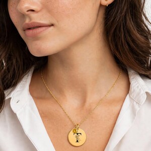 May include: A gold-toned necklace featuring a circular pendant with a black letter T and a small floral design, accompanied by a tiny yellow gemstone charm. The pendant hangs from a delicate gold-toned chain, resting against a white collared shirt.