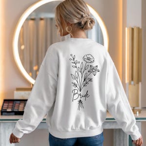 以下が含まれることがあります： 白いスウェットシャツに、黒い花の模様と「Bride」の文字が背中にプリントされています。