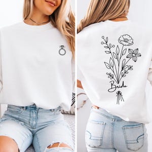 以下が含まれることがあります： 前面にシンプルなリングのデザイン、後ろに花束と「Bride」の文字が書かれた白いスウェットシャツです。