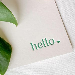 May include: A white card with the word "hello" written in green lettering with a small green heart next to it.