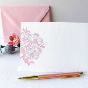 May include: A white card with a pink floral design and a matching pink envelope. A pink and gold pen is on the surface. A decorative flower and a grey ball are also present.