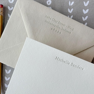 May include: A cream-colored stationery set featuring a note card embossed with the name Michelle Sanders and a matching envelope embossed with the address 2964 Clay Street - No.5, Indianapolis, Indiana 46254, accompanied by a yellow pencil.