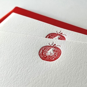 May include: Close-up of white stationery with a red tomato design. The tomato is a detailed illustration, and the stationery has a red border. The paper has a textured surface.