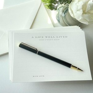 May include: A stack of white rectangular memory cards featuring the printed text A LIFE WELL LIVED, SHARE A FAVORITE MEMORY, and WITH LOVE, accompanied by a black and gold pen. The cards are lined and paired with a matching white envelope.