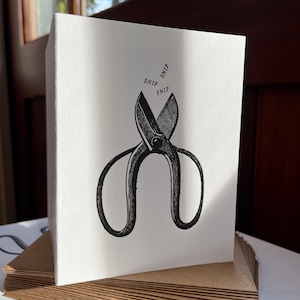 May include: A white card with a black and white illustration of a pair of scissors. The text "SNIP SNIP SNIP" is printed around the scissors.
