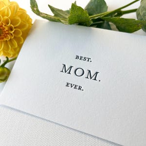 May include: A white card with the words "BEST. MOM. EVER." in a black, serif font. A yellow flower and green leaves are in the background. The card is likely a Mother's Day or birthday card.