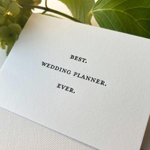 May include: A white card with black text that reads "BEST. WEDDING PLANNER. EVER."