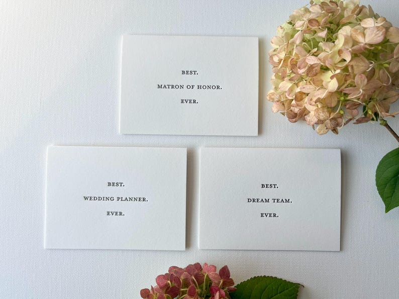 May include: Three white cards with black text. The top card says "BEST. MATRON OF HONOR. EVER.". The middle card says "BEST. WEDDING PLANNER. EVER.". The bottom card says "BEST. DREAM TEAM. EVER."