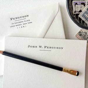 May include: A white envelope with a black return address label that reads "FERGUSON, 15 RAILROAD PLACE, LONG BEACH, NEW YORK 11561". A white card with a black line and the text "JOHN W. FERGUSON" is on top of the envelope. A black pencil is lying on top of the card.