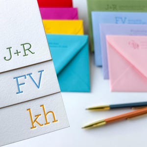 May include: Three white stationery cards featuring embossed monograms in green, blue, and orange. The cards display the text J+R, FV, and kh. In the background, there are several colorful envelopes and two ballpoint pens with gold accents.