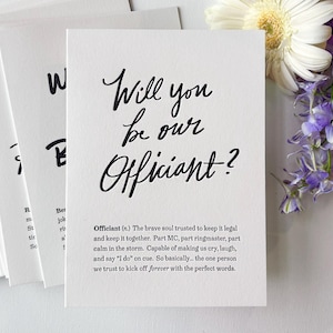 May include: White card with the text "Will you be our Officiant?" in elegant black calligraphy. The card is part of a set, with other cards visible. A bouquet of white and purple flowers is in the background.