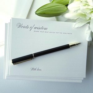 May include: A stack of white rectangular advice cards featuring the black printed text Words of wisdom, SHARE YOUR BEST ADVICE FOR THE NEWLYWEDS, and With love. A black ballpoint pen with gold accents rests diagonally across the top card.