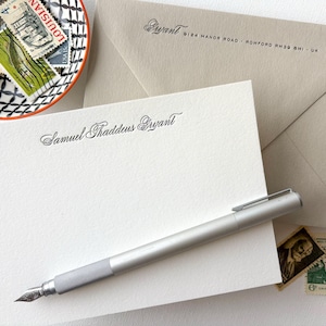 May include: A white stationery card with the name "Samuel Thaddeus Bryant" written in cursive script. A silver pen is lying on the card. The card is on top of a brown envelope with the address "8124 Manor Road, Romford RM3D 8HI, UK" printed on it.