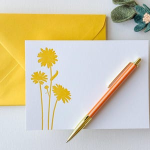 May include: A white card with a yellow envelope. The card has a yellow silhouette of three flowers with stems. A pen with an orange body and gold accents is lying on the card.