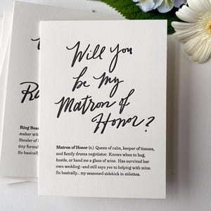 May include: White card with the handwritten text "Will you be my Matron of Honor?" in black script. The card includes a definition of "Matron of Honor" and is part of a set of cards. A white flower and greenery are in the background.