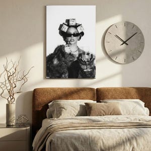 May include: Black and white wall art print of a woman wearing sunglasses, a fur coat, and money curlers in her hair, holding a glass of wine and a cigarette. A round clock hangs on the wall above a bed with neutral-toned bedding.