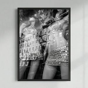 May include: Black and white photograph of two mannequins in a shop window. The mannequins are wearing embellished dresses and skirts. The background shows a city street with buildings and trees.